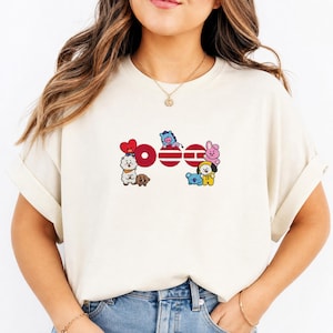 Op de afbeelding: Een crèmekleurig T-shirt met korte mouwen en een grafische print van acht kleurrijke stripfiguren gerangschikt rond drie rode cirkelvormige en horizontale balkvormen. De figuren zijn onder meer een rood hart, een witte lama, een bruine beer, een blauw paard, een blauwe koala, een gele hond en een roze konijn.