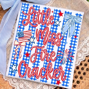 May include: A rectangular sign with a blue and white gingham background featuring the red text Little Miss Fire Cracker. The design includes a small American flag, a light blue bow, red and white fireworks, and several red and white stars.