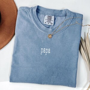 May include: A blue Comfort Colors t-shirt with the word "papa" embroidered in white. A gold necklace with a pendant is draped across the shirt. A brown hat and dried leaves are in the background.