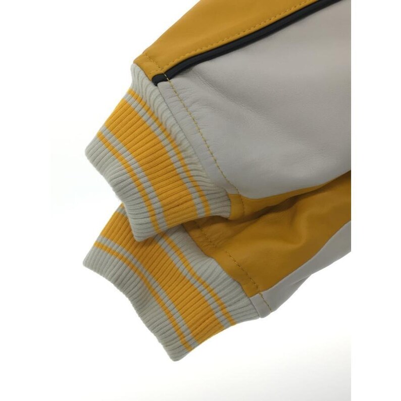 May include: Close-up of a varsity jacket sleeve. The jacket features a white body with yellow accents and a black stripe. The ribbed cuffs are yellow and white striped.