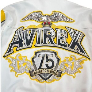 May include: White leather jacket with embroidered details. Features the word "AVIREX" in black and yellow, a shield with "75", and an eagle emblem. The text "AVIATOR'S KING" is also present.