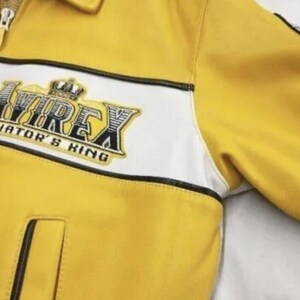 May include: Yellow leather jacket with white and black accents. The jacket features a zipper closure and a patch with the text "AVIREX AVIATOR'S KING". The jacket has a classic design.