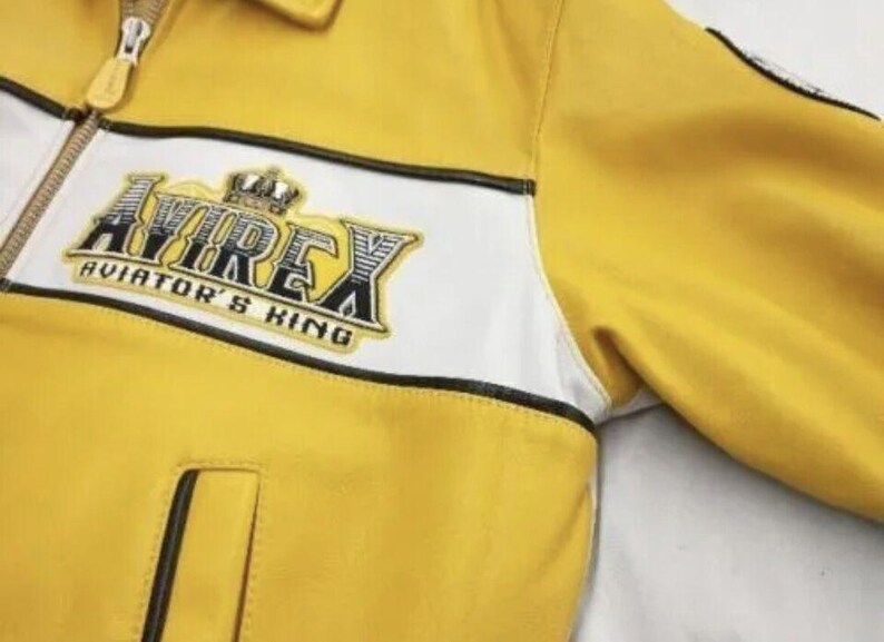 May include: Yellow leather jacket with white and black accents. The jacket features a zipper closure and a patch with the text "AVIREX AVIATOR'S KING". The jacket has a classic design.