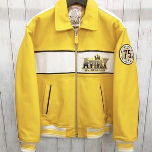 May include: A yellow leather jacket with white stripes and the text "Avirex Aviator's King". The jacket has a collar, zipper, and two front pockets. A patch with the number "75" is on the sleeve.