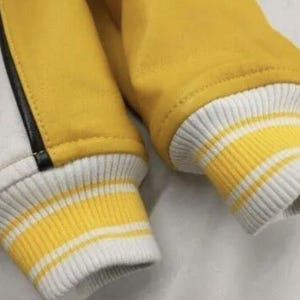 May include: Close-up of a yellow and white varsity jacket. The jacket features yellow sleeves and a white body with a black stripe. The cuffs are ribbed with yellow and white stripes.