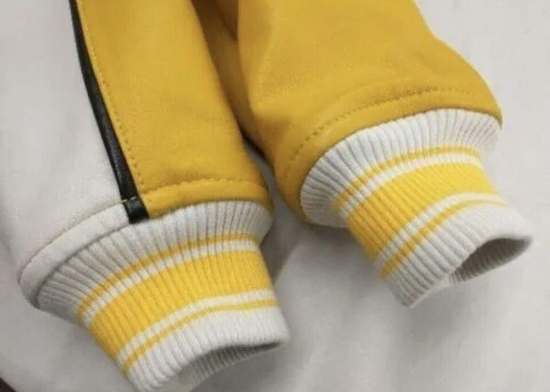 May include: Close-up of a yellow and white varsity jacket. The jacket features yellow sleeves and a white body with a black stripe. The cuffs are ribbed with yellow and white stripes.