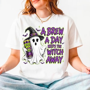 May include: A white t-shirt featuring a graphic of a ghost wearing a black witch hat and holding a purple energy drink can. The text reads A BREW A DAY KEEPS THE WITCH AWAY in purple and green letters, surrounded by small purple bat illustrations.