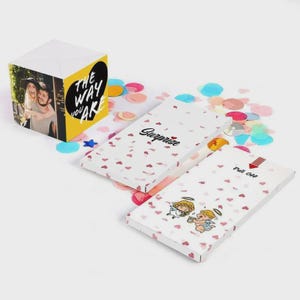 May include: A white gift box with a photo and the text "THE WAY YOU ARE" next to two white boxes with the text "Surprise" and "Pull OFF". The boxes are decorated with red hearts and angel illustrations, with colorful confetti scattered around.