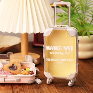 May include: A clear, rolling suitcase-shaped container with the text "HANGOVER recovery kit" and "JESSICA'S SUITCASE" in white. The container has a yellow interior and is on a wooden surface. A smaller pink suitcase is in the foreground.