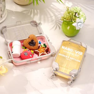 Puede incluir: Un pequeño recipiente transparente en forma de maleta con el texto "HANG OVER recovery kit" y "JESSICA'S SUITCASE". El recipiente es amarillo con detalles grises. En el interior, un recipiente rosa contiene golosinas y una botella blanca. También se ve un segundo recipiente abierto.