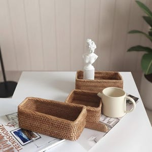 May include: Three rectangular woven rattan baskets of varying sizes are arranged on a white surface, alongside a small white bust sculpture and a cream-colored mug. The baskets are a warm brown color.