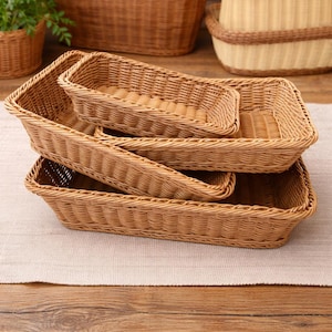 May include: A stack of four rectangular woven baskets in varying sizes. The baskets are made of light brown wicker and have a textured surface. The baskets are empty and are sitting on a light-colored surface.