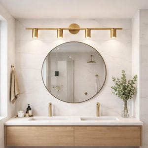 May include: A modern bathroom vanity with a round mirror, gold fixtures, and a four-light vanity light fixture. The vanity has a light wood finish with two drawers. A vase of greenery sits on the countertop.