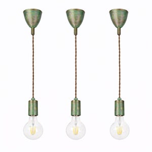 Puede incluir: Tres lámparas colgantes con estética vintage. Cada lámpara presenta una pantalla y base de metal en tono verde, un cable trenzado marrón y una bombilla redonda transparente. Las lámparas están suspendidas sobre un fondo blanco.