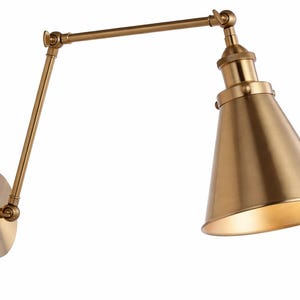 Puede incluir: Una lámpara de pared de color latón con un brazo ajustable y una pantalla cónica. La lámpara tiene una base circular y un brazo giratorio para una iluminación direccional. La pantalla está inclinada hacia abajo, con un interior dorado más claro.