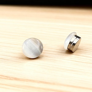 Puede incluir: Dos pendientes redondos de botón de piedra ojo de gato gris. Un pendiente está en posición vertical y el otro está de lado, revelando la parte trasera de metal y el poste de plástico blanco. Sobre una superficie de madera clara.