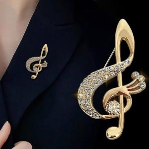 Może przedstawiać: Złota broszka w kształcie klucza wiolinowego, ozdobiona błyszczącymi, przezroczystymi kryształami. Przypinka z nutą muzyczną jest prezentowana na ciemnym tle, podkreślając jej elegancki design. Widoczna jest również mniejsza wersja broszki.