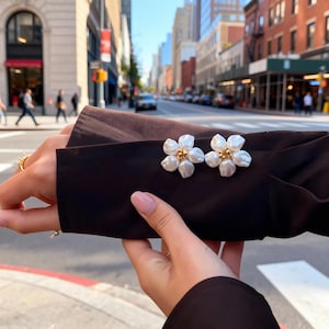 May include: A pair of flower-shaped earrings with white petals and gold-colored centers. The earrings are displayed on a dark brown jacket sleeve. The background shows a city street with buildings and pedestrians.
