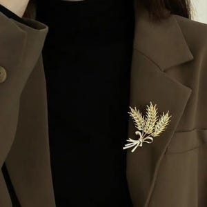 Può includere: Una spilla color oro a forma di spighe di grano e fiocco, appuntata su un blazer marrone. La spilla è rifinita con piccoli dettagli scintillanti. Il blazer è indossato sopra un top nero.