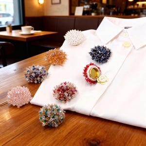 May include: A collection of beaded brooches in various colors, including pink, orange, blue, and red, displayed on a white shirt. The brooches feature intricate designs with small, sparkling beads, and some have gold-colored metal accents.