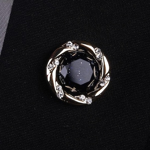 Puede incluir: Un botón dorado con una piedra central facetada de color azul oscuro. El botón está rodeado por un diseño dorado retorcido, acentuado con pequeños cristales transparentes. El botón está sobre un fondo de tela negra.