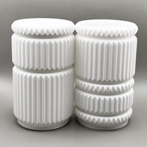 May include: Two white cylindrical objects with vertical fluted textures and horizontal beaded bands. The left object features a single fluted section, while the right object displays two fluted sections separated by a central beaded ring.