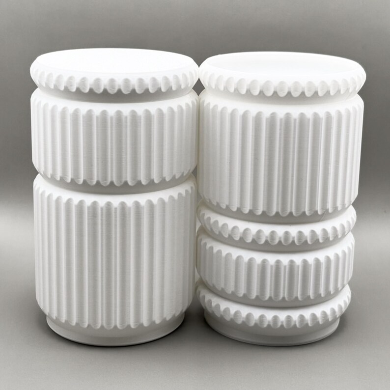May include: Two white cylindrical objects with vertical fluted textures and horizontal beaded bands. The left object features a single fluted section, while the right object displays two fluted sections separated by a central beaded ring.