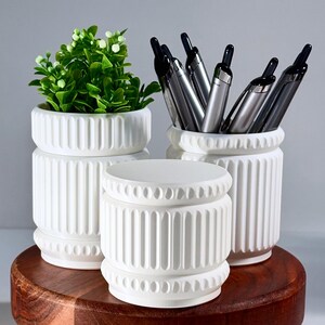 Puede incluir: Tres recipientes cilíndricos blancos con texturas acanaladas verticales y bordes con cuentas. Un recipiente contiene una planta artificial verde, otro contiene varios bolígrafos plateados y negros, y el tercero está vacío en el centro.