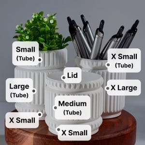May include: A collection of white cylindrical containers with vertical ribbed textures, labeled as Small (Tube), Large (Tube), X Small (Tube), X Small, Medium (Tube), Lid, and X Large. Some containers hold green plants or silver pens.