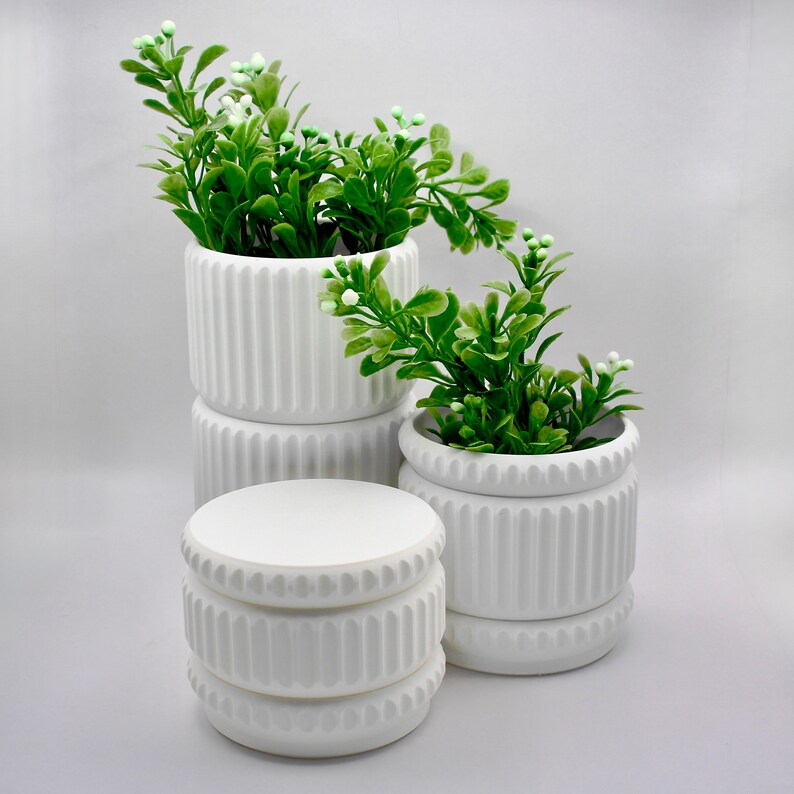 May include: Three white ceramic planters with a vertical ribbed texture. Two of the planters contain artificial green leafy plants with small white buds. One planter is empty and sits in the foreground, featuring a matching ribbed design.