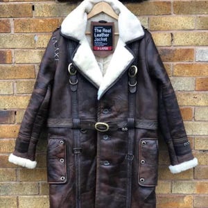 May include: A brown leather jacket with a white shearling collar and lining. The jacket features a belt, multiple buckles, and a button-down front. The label reads "The Real Leather Jacket" and "X-LARGE".
