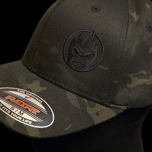 Peut inclure: Casquette de baseball Flexfit camouflage noir et vert avec un logo brodé noir. La casquette a une visière incurvée et le texte "THE ORIGINAL FLEXFIT S/M IT FITS YOUR LIFE" sur un autocollant.