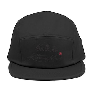 Op de afbeelding: Zwarte vijf-panel cap met een platte klep. De cap heeft Japanse karakters en de tekst "Kitsura Mori" in donkerrood. Een kleine rode cirkel is ook zichtbaar aan de rechterkant van de cap.