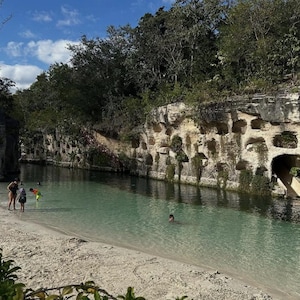 May include: A scenic view of a natural swimming hole with clear turquoise water. The water is surrounded by limestone cliffs with lush green vegetation. Several people are enjoying the water and the sandy beach.