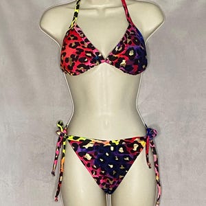 May include: A two-piece bikini set with a vibrant leopard print. The top is a halter style, and the bottoms are tie-side. The print features a mix of red, pink, purple, and gold, creating a bold and eye-catching look. The bikini is displayed on a mannequin.
