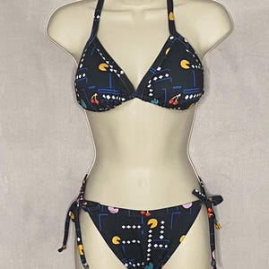May include: Black bikini set with a Pac-Man video game pattern. The halter top and tie-side bottoms feature a repeating design of the iconic game's characters and elements in white, yellow, blue, and red. The bikini is displayed on a mannequin.