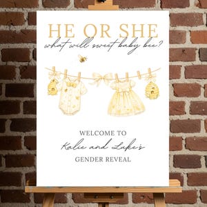 May include: A gender reveal sign with the text "HE OR SHE what will sweet baby bee?" in gold lettering. The sign features illustrations of baby clothes and a bee. The sign welcomes guests to Kalie and Luke's gender reveal.