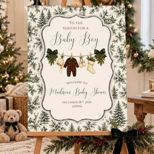 K&ouml;nnte beinhalten: Ein rechteckiges Schild mit cremefarbenem Hintergrund und dunkelgr&uuml;nem botanischem Rand. Der Text lautet 'Tis the season for a Baby Boy', 'Welcome to Madison's Baby Shower', 'December 18th, 2026' und '2:00pm'. Es zeigt eine W&auml;scheleine mit einem Strampler.