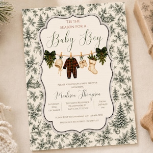 K&ouml;nnte beinhalten: Eine rechteckige Einladungskarte f&uuml;r eine Babyparty mit cremefarbenem Hintergrund und einem dunkelgr&uuml;nen botanischen Muster aus Tannen und Stechpalmen. Der Text lautet 'Tis the season for a baby boy' &uuml;ber einer W&auml;scheleine mit Babykleidung.