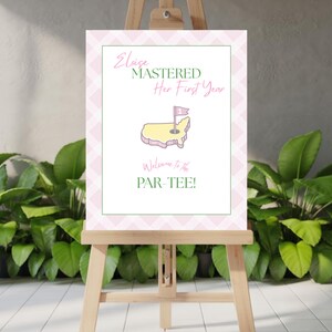 Könnte beinhalten: Ein Schild auf einer hölzernen Staffelei mit dem Text "Eloise Mastered Her First Year" und "Welcome to the PAR-TEE!" in Grün und Rosa. Das Schild zeigt eine gelb-rosa Umrisszeichnung eines Golfplatzes mit einer Fahne. Der Hintergrund hat ein rosa-weißes Rautenmuster.