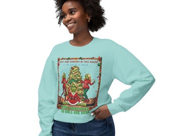 Può includere: Una felpa a maniche lunghe azzurra con una grafica del Grinch e altri personaggi simili al Grinch attorno a un albero di Natale. Il testo sulla felpa recita: "HERE'S SOME GRINCHES IN THIS HOUSE IT THE QUEENS OF MEAN. MERRY GRINCHMAS THE GIRLS ARE HERE."
