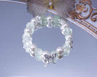 May include: A beaded bracelet featuring a mix of white pearls and pale green beads. A silver butterfly charm hangs at the center. The bracelet is displayed on a reflective surface, with decorative elements in the background.