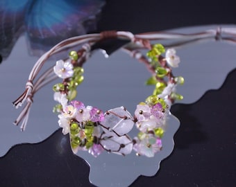 May include: A floral bracelet with white, pink, and green beads. The bracelet is made with a brown and white cord and features a cluster of small, delicate flowers. The bracelet is displayed on a reflective surface.