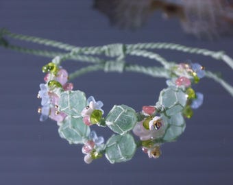 May include: A delicate bracelet featuring light green, faceted beads encased in a woven net, interspersed with small pink, blue, and green floral accents. The bracelet is adjustable with a light green cord.
