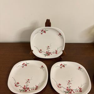 Puede incluir: Juego de tres platos blancos cuadrados con esquinas redondeadas. Cada plato presenta un diseño floral con flores rojas y tallos verdes. Un plato se exhibe en un soporte de madera.