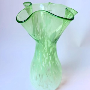 Op de afbeelding: Een lichtgroene glazen vaas met een uitlopende, gegolfde rand en een taps toelopende hals die uitloopt in een ronde voet. Het glas vertoont een doorschijnend, gemarmerd wit en lichtgroen patroon over het hele lichaam voor een zacht effect.