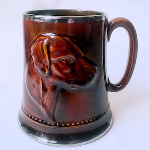 May include: A brown ceramic mug featuring a raised, three-dimensional relief of a dog's head on the front. The mug has a glossy finish, a curved handle, and a metallic silver-toned rim and base, with a small beaded detail above the base.