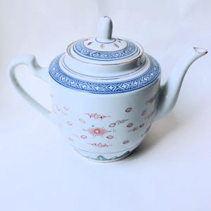 May include: A white porcelain teapot featuring a blue geometric border pattern around the lid and upper rim. The body is decorated with small red and grey floral motifs and a central sunburst design. It includes a matching lid with a knob.