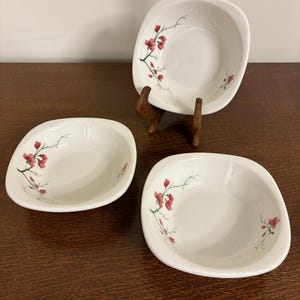 May include: Three white, square-shaped ceramic bowls with rounded corners. Each bowl features a delicate floral design in red and green. One bowl is displayed on a wooden stand. The bowls are arranged on a dark brown surface.