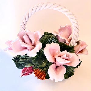 May include: A ceramic basket featuring a twisted white handle and a decorative arrangement of glossy light pink roses with dark green leaves. The base of the floral cluster includes a small dark red bud and a hint of orange-toned foliage at the bottom.